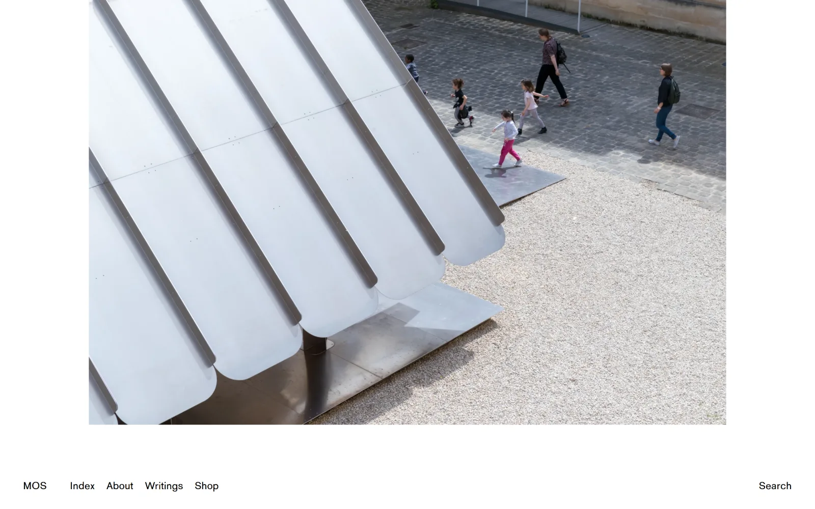 mos.nyc, architect website design: a photograph of a white fluted canopy on gravel, people crossing cobbles behind it